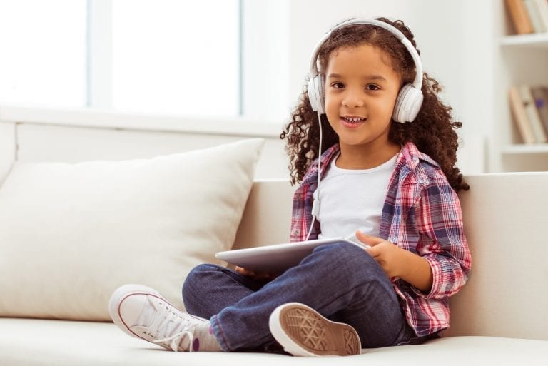 Cute little Afro-American girl in casual clothes and headphones using a tablet and listening to music while sitting on a sofa in the room.